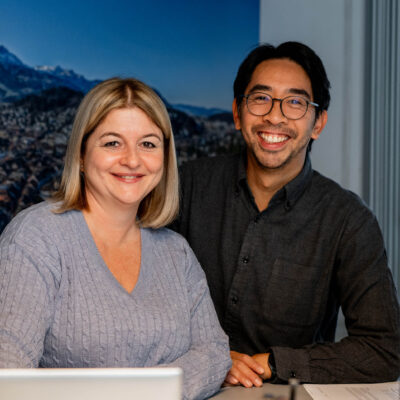 Zwei Mitarbeitende von Röösli steht hinter einem Arbeitstisch und lachen in die Kamera. Röösli Funktionsdecken.